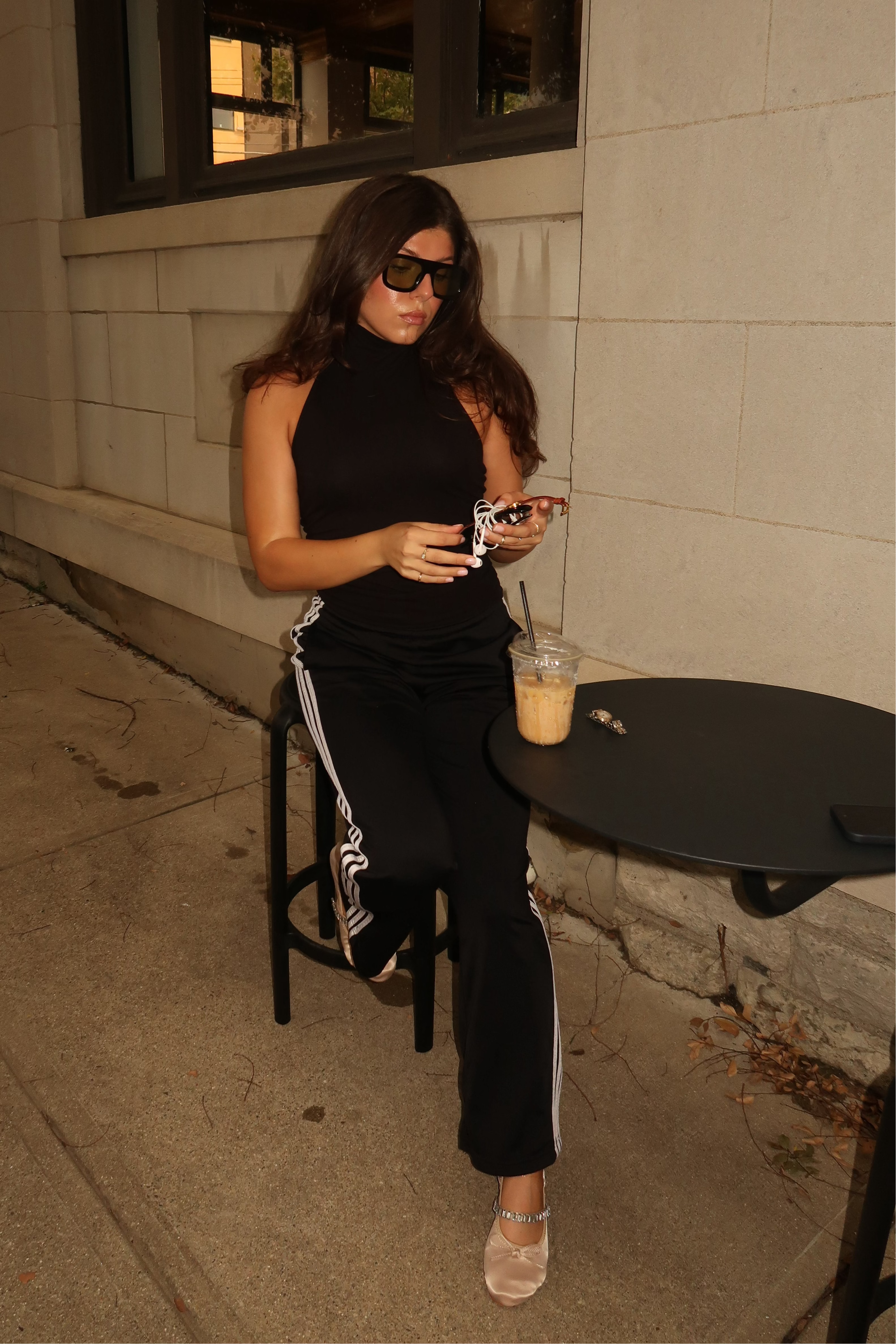 A woman wearing a black sleeveless mock-neck top and black track pants with white side stripes sits on a black stool at an outdoor cafe table, looking down at her phone. Creative Direction Branding Agency start up inspiation g7x women coffee cute outfit ballet flats